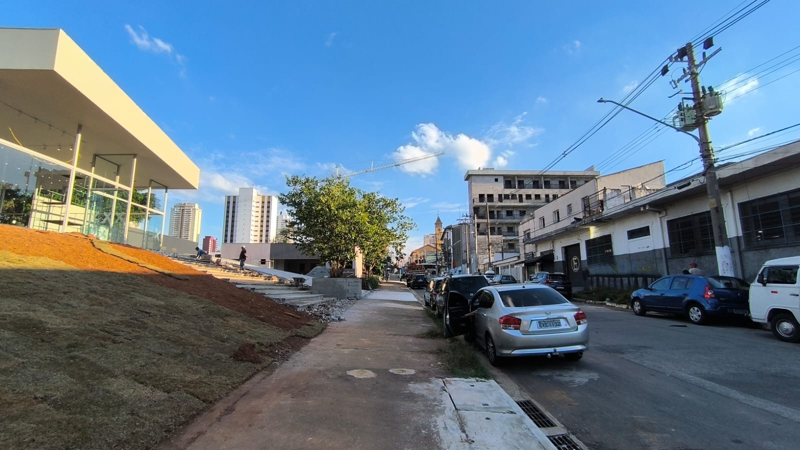 Vista da rua do stand de vendas do Orbya Vila Carrão em construção, com fachada de vidro e paisagismo recém-instalado na Av. Conselheiro Carrão