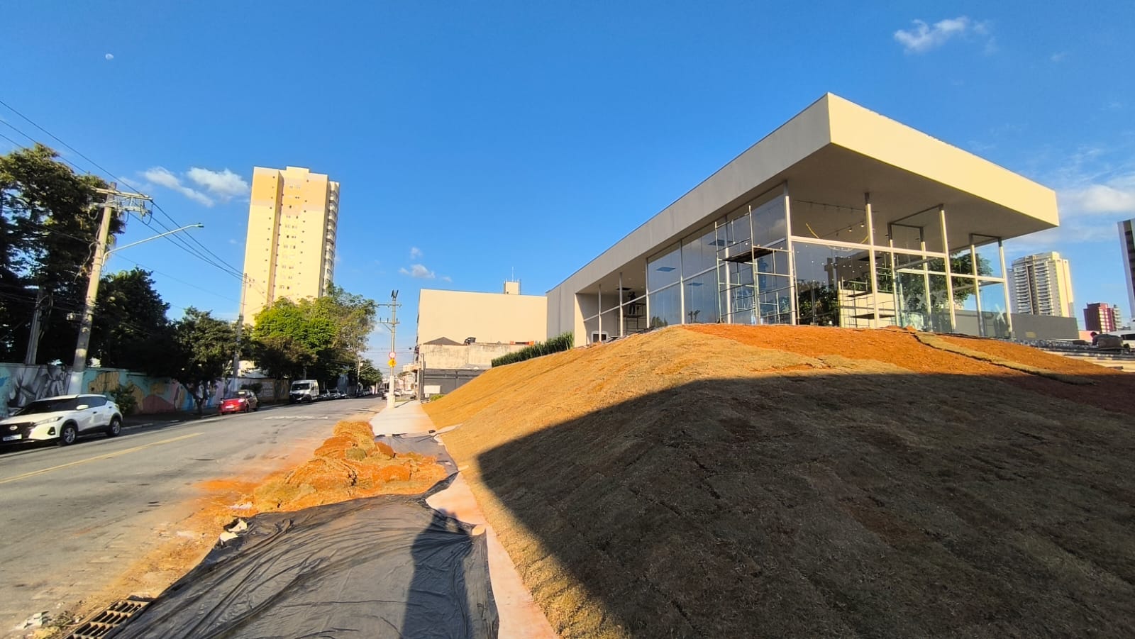 Stand de vendas do Orbya Vila Carrão visto da esquina, com gramado recém-plantado e torre residencial ao fundo na Vila Carrão